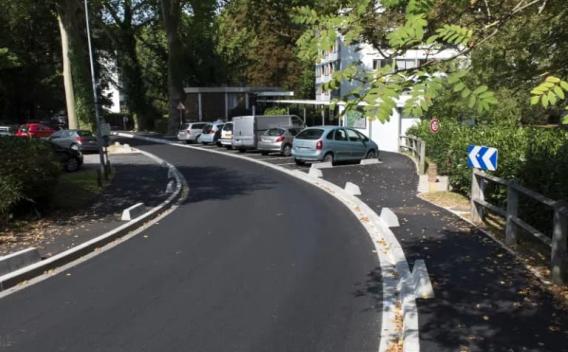 Voirie urbaine rénovée sous couvert boisé : enrobé noir lisse, caniveaux et bordures béton neufs, stationnements latéraux et giratoire à l’arrière-plan.