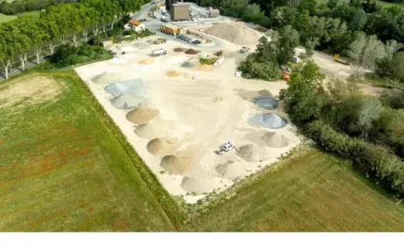 Plateforme de négoce de granulats vue aérienne, stocks en cônes de sables et graviers, engins de manutention au bord de champs et d’un bois.