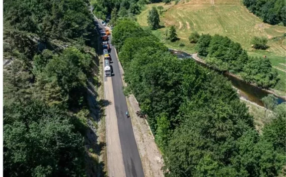 Chantier de réfection de chaussée en milieu rural : ruban routier neuf bordé de forêt et de prairies, convoi d’engins et de camions en file.