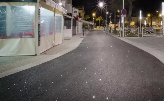Voie urbaine piétonne revêtue d’un enrobé noir lisse (pose récente) bordée de commerces, éclairage public nocturne et mobilier urbain.