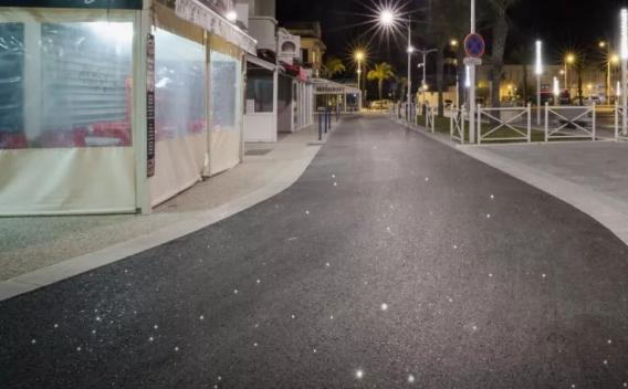 Voie urbaine piétonne revêtue d’un enrobé noir lisse (pose récente) bordée de commerces, éclairage public nocturne et mobilier urbain.