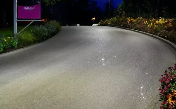 Route en courbe éclairée la nuit, bordée de massifs fleuris et d'une affiche, surface propre et brillante.