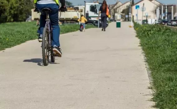 Allée piétonne/cyclable fraîchement revêtue en milieu naturel et urbain : surface claire et lisse, bordée d’herbe et d’arbres, avec un banc en bois au bord et des usagers (piétons et cyclistes) en circulation.
