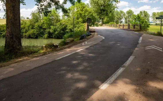 Un tronçon de route bordant une rivière, léger virage vers un petit pont, ombrage d’arbres, marquage au sol blanc, ambiance champêtre et lumière douce.