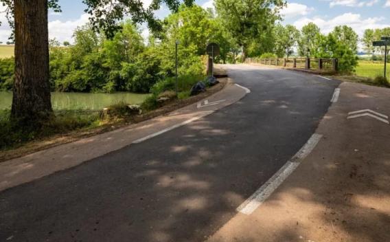 Un tronçon de route bordant une rivière, léger virage vers un petit pont, ombrage d’arbres, marquage au sol blanc, ambiance champêtre et lumière douce.