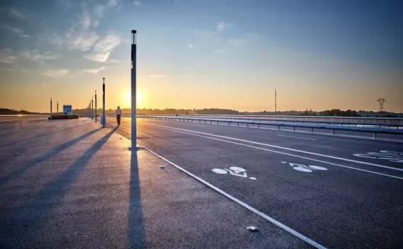 Aire de stationnement neuve au lever du soleil, enrobés fraîchement posés avec marquages au sol (zones piétons et cycles) et rangée de mâts d’éclairage projetant de longues ombres.