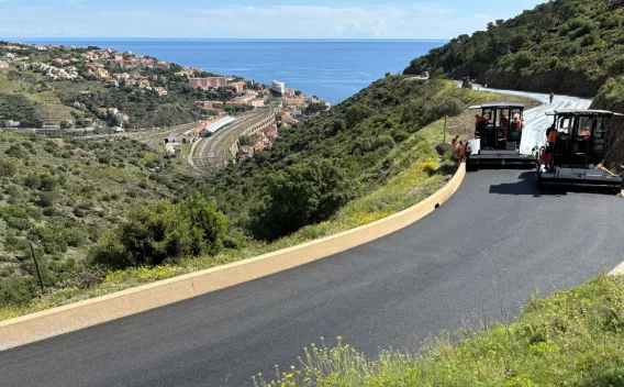 Revêtement routier neuf sur route de montagne surplombant le littoral, avec deux finisseuses asphaltant une voie sinueuse, vue sur village et lignes ferroviaires en contrebas.