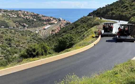 Revêtement routier neuf sur route de montagne surplombant le littoral, avec deux finisseuses asphaltant une voie sinueuse, vue sur village et lignes ferroviaires en contrebas.