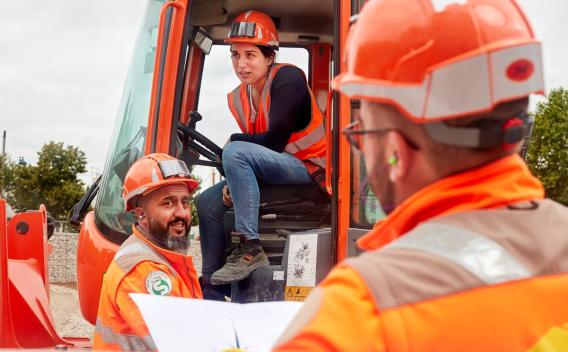 Équipe de chantier en gilets haute visibilité et casques échangeant des consignes autour d’une pelle mécanique.