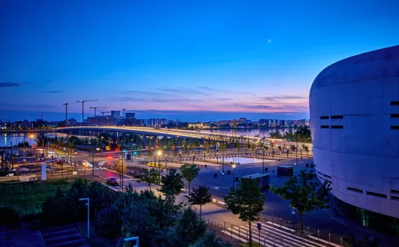 Création du tablier du pont Simone Veil à Bordeaux par Colas : Vue nocturne d’un aménagement urbain riverain éclairé : pont, voies piétonnes et espace public aménagé le long de l’eau.