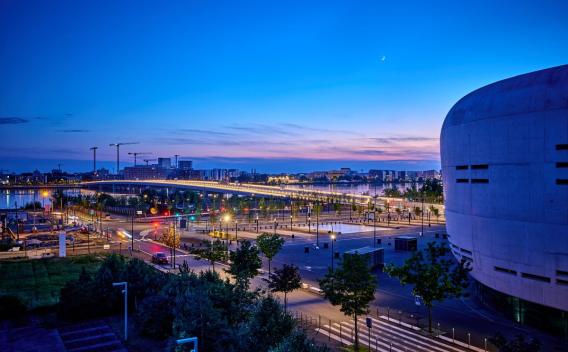Création du tablier du pont Simone Veil à Bordeaux par Colas : Vue nocturne d’un aménagement urbain riverain éclairé : pont, voies piétonnes et espace public aménagé le long de l’eau.