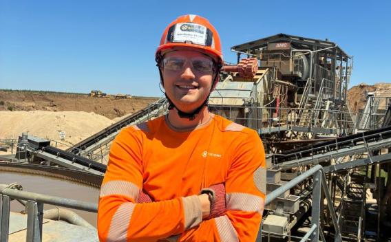 Collaborateur portant un casque et des équipements de protection, debout devant une installation industrielle de traitement des matériaux sous un ciel bleu.