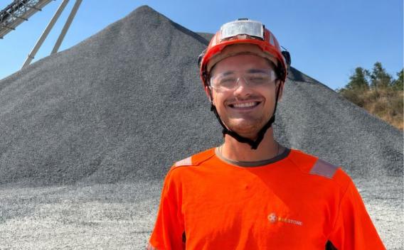 Collaborateur souriant en tenue de sécurité orange et casque, devant un important stock de granulats sur un site industriel.