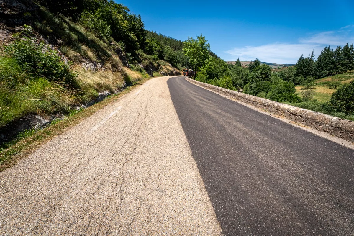 Route de montagne partiellement remise à neuf : une moitié d’enrobé neuf bordée de végétation et d’un talus rocheux, engin de chantier au loin sous un ciel bleu.