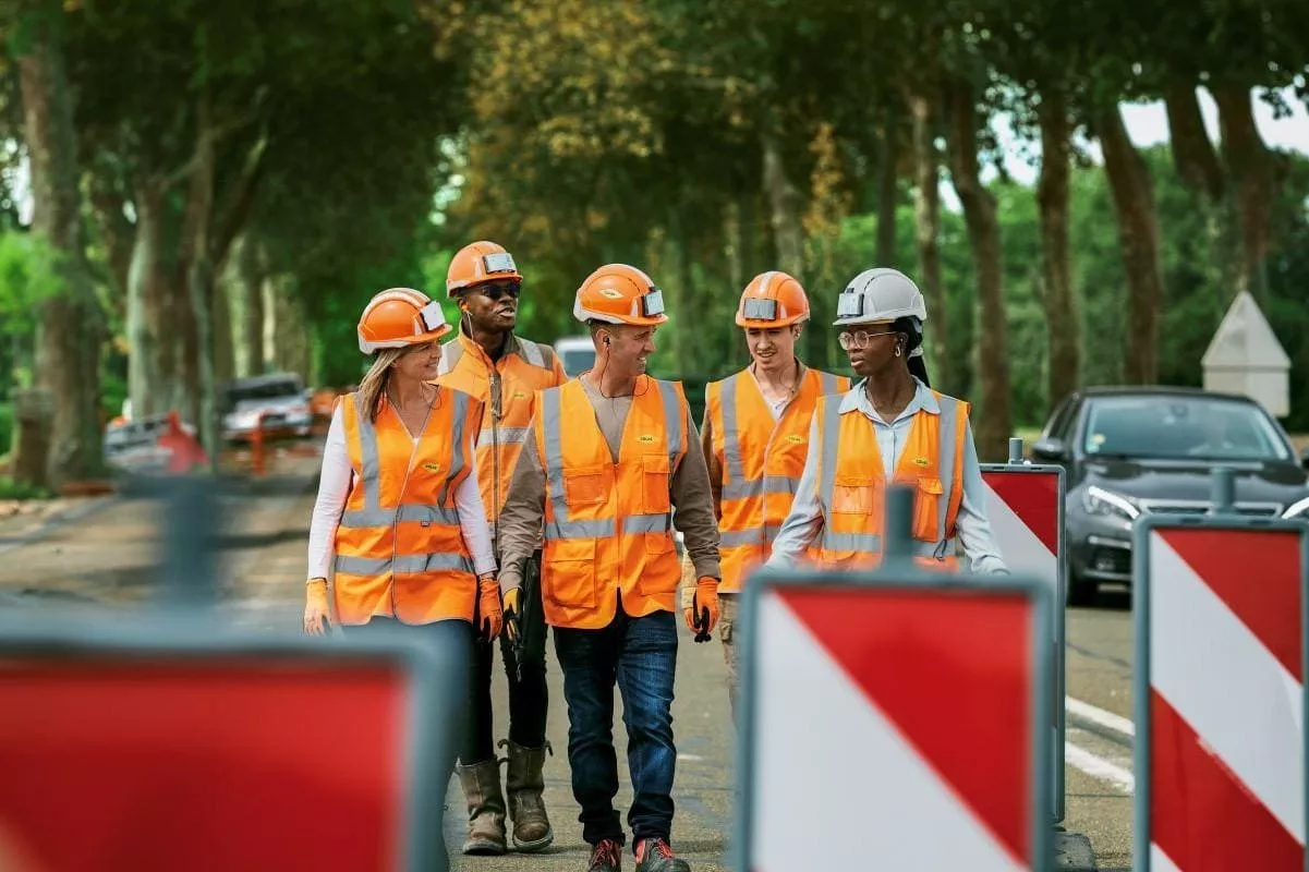 Colas recrute partout en France : Équipe de chantier en tenue de sécurité (gilets haute visibilité et casques) marchant le long d’une chaussée avec balisage et circulation en arrière-plan.