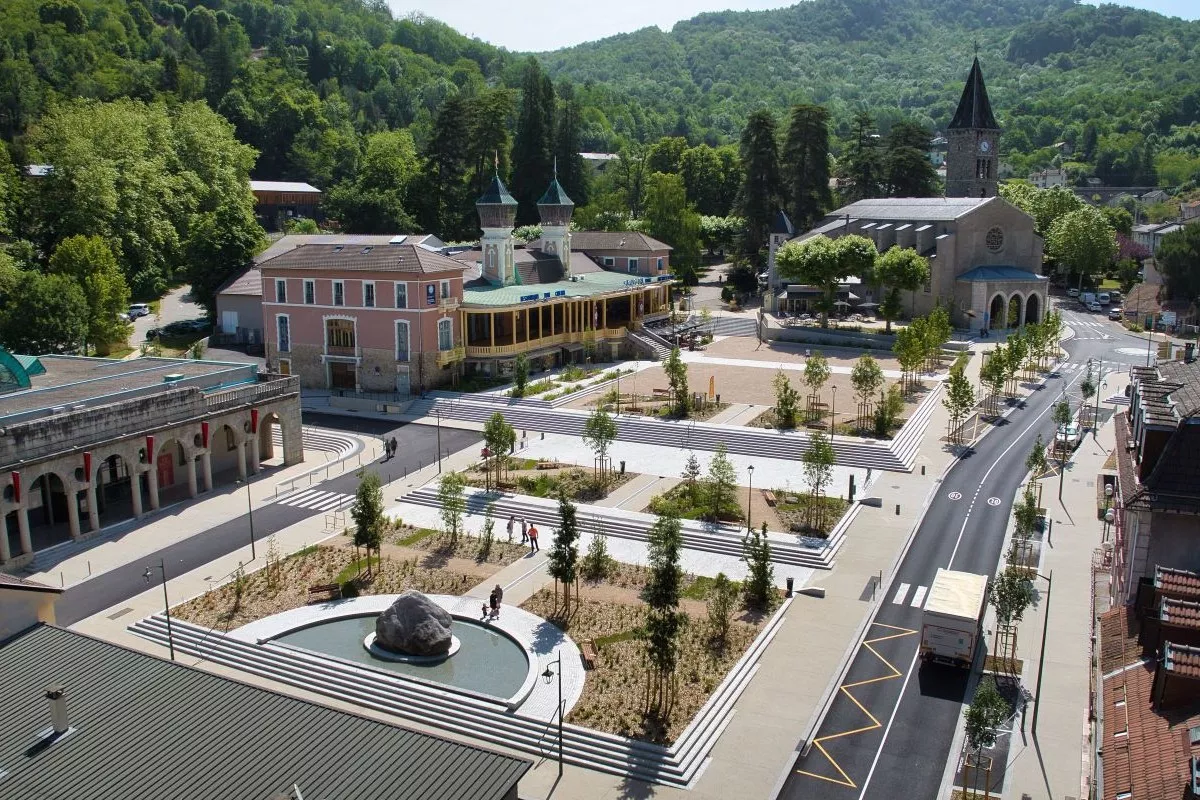 Place publique réaménagée : esplanade plantée avec bassins et gradins, bâtiments historiques en bordure et une rue récemment rénovée.
