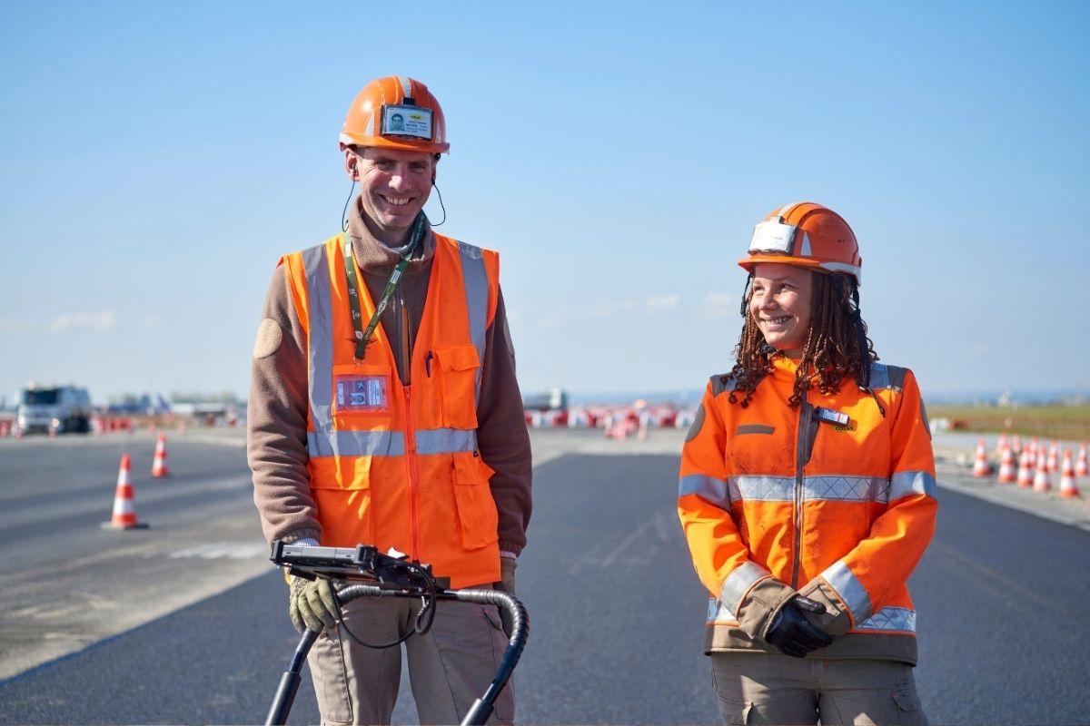 Deux collaborateurs Colas en tenue de chantier orange sur une piste, équipés de casques de sécurité.