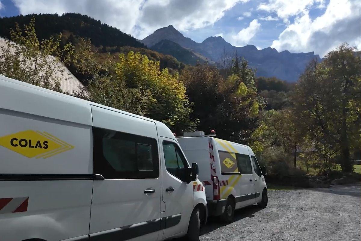 Deux véhicules de chantier Colas stationnés au pied d’un massif montagneux boisé.
