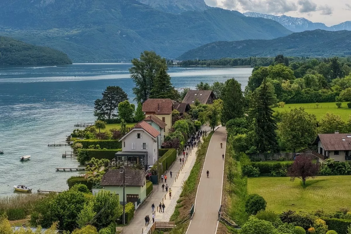 Vue aérienne d’une voie cyclable longeant un lac au pied des montagnes, avec des cyclistes et des piétons circulant dans un environnement verdoyant.