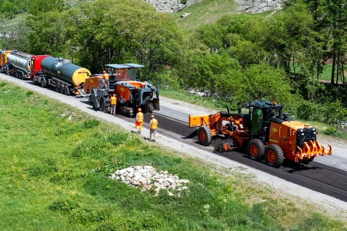 Des équipes de travaux publics réalisent la réfection d’une chaussée avec une finisseuse, une niveleuse et des camions-citernes dans un environnement montagneux.