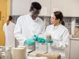 Scientifiques au Core Center : Deux techniciens en blouse blanche dans un laboratoire de matériaux. Ils manipulent des éprouvettes et des granulats de béton sur une table remplie d'échantillons et d'outils de laboratoire ; on voit des contenants, des morceaux de béton et des appareils d'essai en arrière-plan.