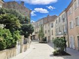 Ruelle piétonne d'un centre‑ville provençal : pavage clair à larges dalles, façades anciennes aux volets pastel, petits massifs plantés et arbustes, quelques boutiques et terrasses, ciel bleu ponctué de nuages blancs.