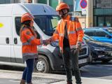 Deux collègues en tenue et casque de chantier s'échangent une poignée de main au bord d'une zone urbaine, fourgon de chantier et véhicules en arrière‑plan.