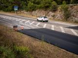 Bande de chaussée fraîchement rechargée en enrobé noir le long d’un échangeur, avec cône de signalisation et véhicule passant à proximité d’un talus boisé.