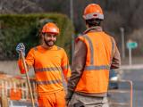 Échange sur zone de chantier routier entre deux opérateurs en EPI orange, l’un tenant un outil de manutention près d’un balisage et d’une voie ouverte à la circulation.