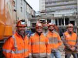 Quatuor d’ouvriers en chantier devant un bâtiment démoli, équipés d’EPI (casques et gilets haute visibilité) et engin de terrassement en arrière-plan, site en phase de déconstruction.