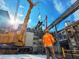 Grutage industriel sur une plateforme en milieu d'usine : grande grue mobile levant une structure métallique, opérateur en tenue de chantier au premier plan, appareillage et passerelles industrielles en arrière‑plan.