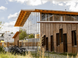 Un bâtiment contemporain en bois et verre, à grande toiture inclinée, se dresse sous un ciel partiellement nuageux avec un parking à vélos au premier plan.