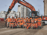 Groupe d'ouvriers en tenue de chantier orange posant devant une pelle hydraulique sur un chantier urbain.
