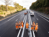 Une équipe de travailleurs en tenue haute visibilité installe une glissière de sécurité sur une autoroute neuve, avec véhicules et matériel de chantier alignés sur la voie.