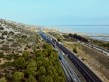 Vue aérienne d’une autoroute côtière à plusieurs voies longeant des étangs, avec trafic soutenu, voie ferrée parallèle et reliefs végétalisés.
