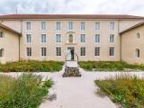 Cour d’édifice historique rénové : façade symétrique enduite couleur sable, toitures tuiles rouges, parterre minéral clair encadré de massifs fleuris et puits central en pierre.