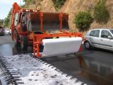 Opération d’enrobage renforcé en cours : un engin routier (chargeuse/finisseur) déroule un géotextile blanc sur une surface d’enrobé frais tandis qu’un film d’enrobé noir est appliqué en fond. Véhicules circulent à proximité, et la zone montre des traces de pneus et d’application récentes.