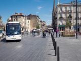 Place urbaine pavée animée : bus, piétons, potelets d’alignement, façades historiques et flèche d’église sous ciel bleu.