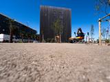 Place urbaine en travaux avec engin de terrassement et plantation d'arbres en cours, bordée d'immeubles et de véhicules de chantier.