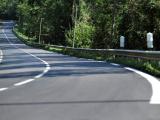 Route départementale sinueuse en lisière de forêt : enrobé neuf au noir profond, marquage blanc contrasté et glissière métallique continue en rive.