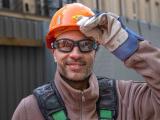 Portrait d’un ouvrier de chantier levant la main gantée vers son casque de sécurité orange, harnais et gilet de protection visibles sur fond de palissade urbaine.