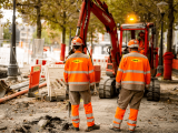 Deux opérateurs en tenue de chantier Colas supervisant une mini-pelle sur un trottoir urbain en cours de rénovation, mettant l’accent sur la coordination et les mesures de sécurité.