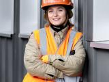 Travailleur en gilet haute visibilité et casque de chantier, appuyé contre une structure métallique, observant le chantier ou en pause entre deux interventions. Équipement conforme : casque de protection, gilet réflecteur, vêtements de chantier et gants dans la main.