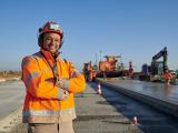 Ouvrier en tenue de sécurité orange sur un chantier routier de pose de chaussée avec engins et équipe au travail en arrière‑plan.