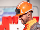 Collaborateur Colas : Ouvrier de chantier en gilet haute visibilité orange et casque, mains sur les hanches, posé sur une promenade urbaine en bord de mer, avec bâtiments et muret en pierre à l’arrière-plan.