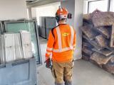 Opérateur en EPI (casque, protections auditives, gants et hauts visibilité) dans un local en réhabilitation, surveillant le stockage de matériaux isolants et menuiseries en attente d’installation.