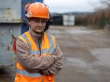 Conductrice d'engins chez Colas : Ouvrière de chantier en EPI Colas (casque, gilet haute visibilité) bras croisés sur une zone industrielle extérieure.