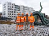 Equipe Colas en charge de travaux : Cinq ouvriers en tenues orange posent sur un parvis pavé devant une sculpture tentaculaire et un immeuble contemporain.