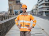 Collaborateur Colas : Ouvrier de chantier en gilet haute visibilité orange et casque, mains sur les hanches, posé sur une promenade urbaine en bord de mer, avec bâtiments et muret en pierre à l’arrière-plan.