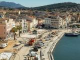 Aménagement urbain en front de mer: réfection des quais et de l’esplanade avec terrassements, pose de bordures et réseaux, engins de chantier et matériaux, bordés par immeubles colorés, port et bateaux, relief montagneux en arrière-plan.